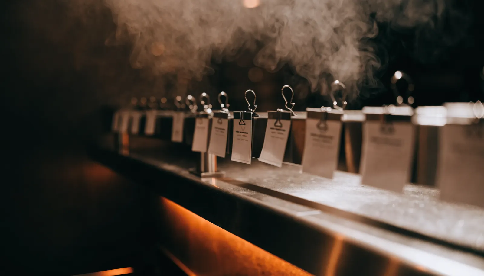 A stainless kitchen pass with blank tickets clipped to the rail, steam rising